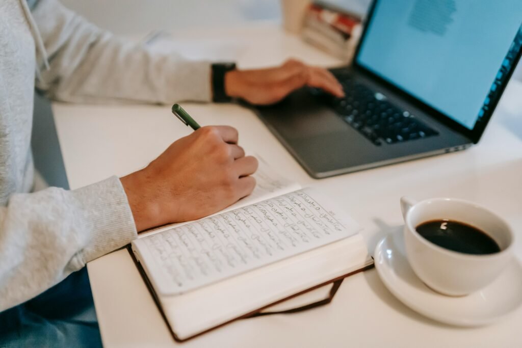 A person crossing off a completed task in a notebook beside a laptop and coffee